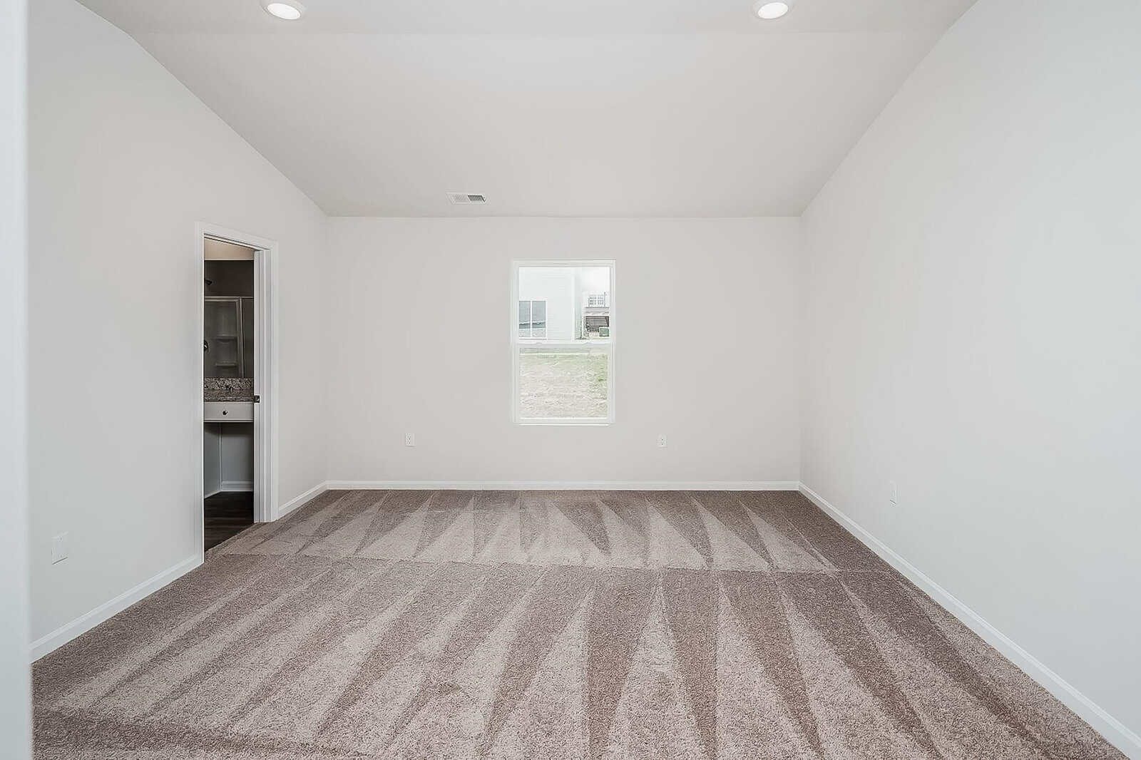 An empty room with beige carpet and white walls. There is a window on the far wall, and an open door leading to another room, possibly a bathroom, on the left. The ceiling is vaulted with recessed lighting.