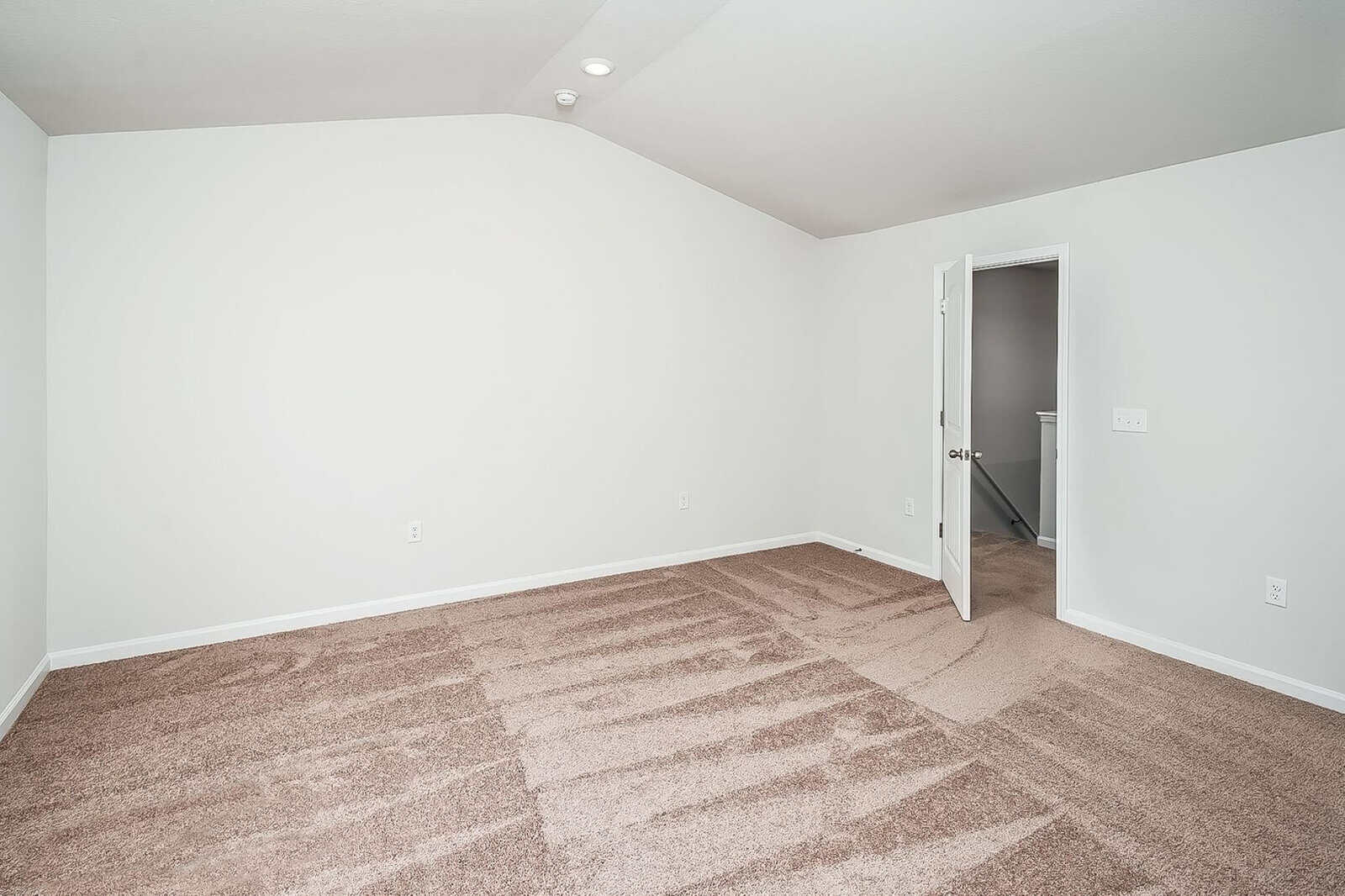 An empty room with white walls, a vaulted ceiling, and beige carpet. The room has an open door leading to a hallway. There are no furnishings or decorations.