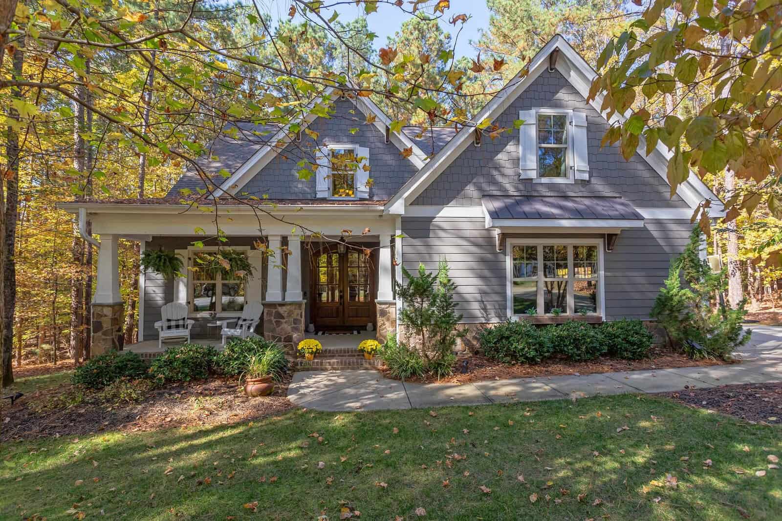 A gray house with white trim and a front porch sits amid tall trees and autumn foliage at 3587 Cotton Field Court, Wake Forest. The yard features green grass, potted flowers, and bushes lining the walkway to a wooden front door.