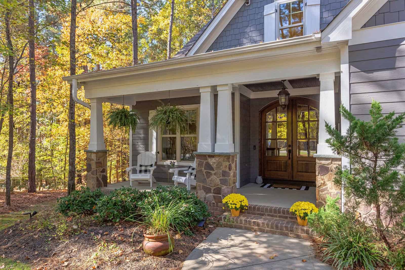 A cozy house with gray siding, stone accents, and a front porch featuring white chairs, hanging ferns, and yellow potted flowers, surrounded by trees and greenery in a wooded setting.