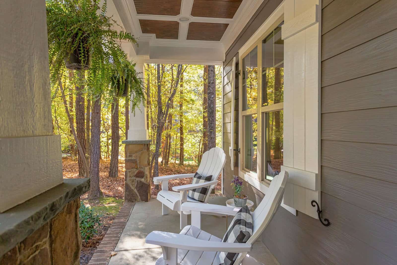 A cozy front porch with two white Adirondack chairs, a small table, a hanging fern, and a wooded landscape in the background on a sunny day.