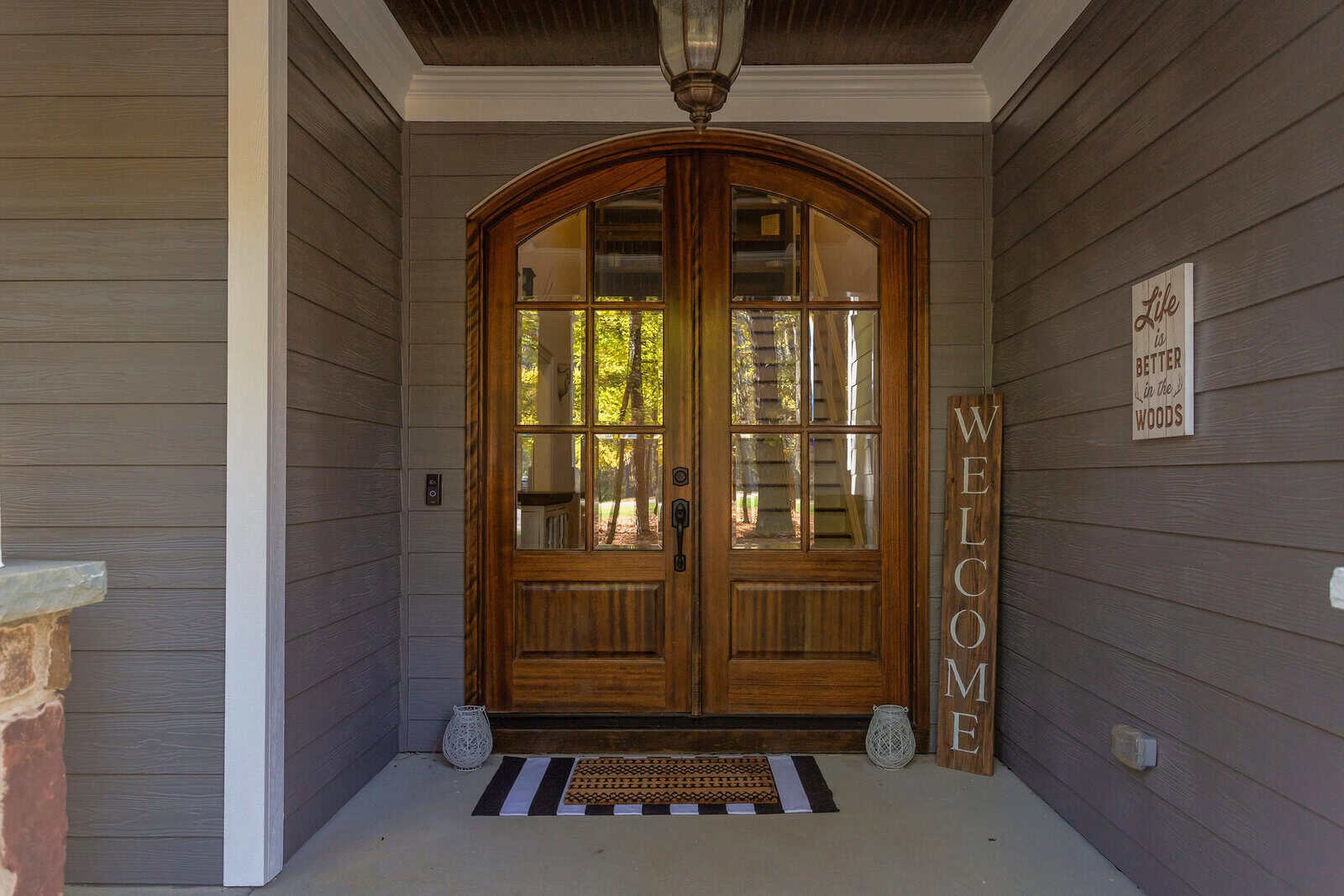 A wooden double front door with glass panels is framed by gray siding. A vertical "WELCOME" sign and a mat decorate the entrance. A wall sign reads "Life is better in the woods," and trees are visible through the glass.