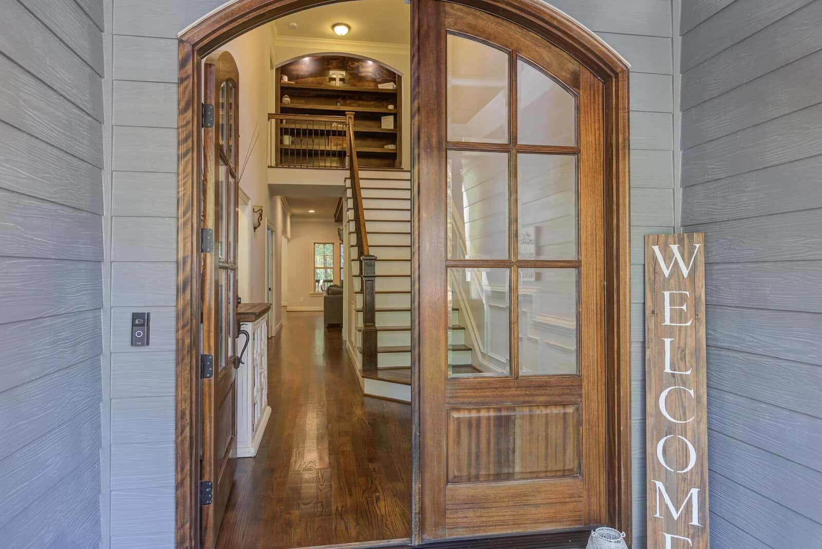 A welcoming entryway with a wooden front door, a "WELCOME" sign, hardwood floors, white walls, staircase, and a view into a well-lit, modern home interior.
