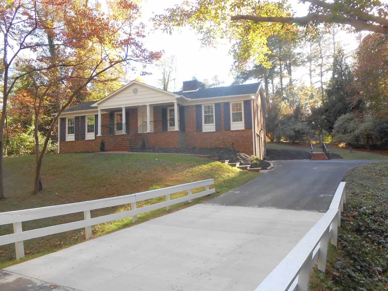 A brick single-story house at 227 Willowood Drive, Henderson, NC with white trim, surrounded by trees with autumn foliage. A white fence runs along the driveway leading to the house.