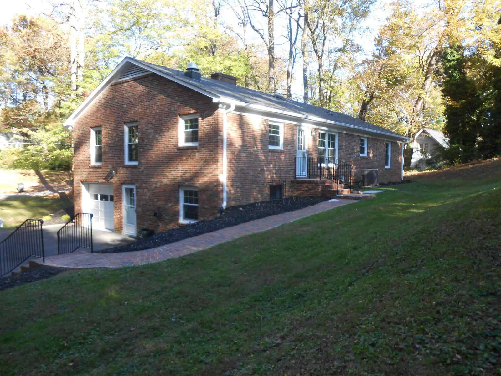A brick single-story house with white trim and a black roof, surrounded by a green lawn on a sunny day. a paved path leads to the inclined entrance.