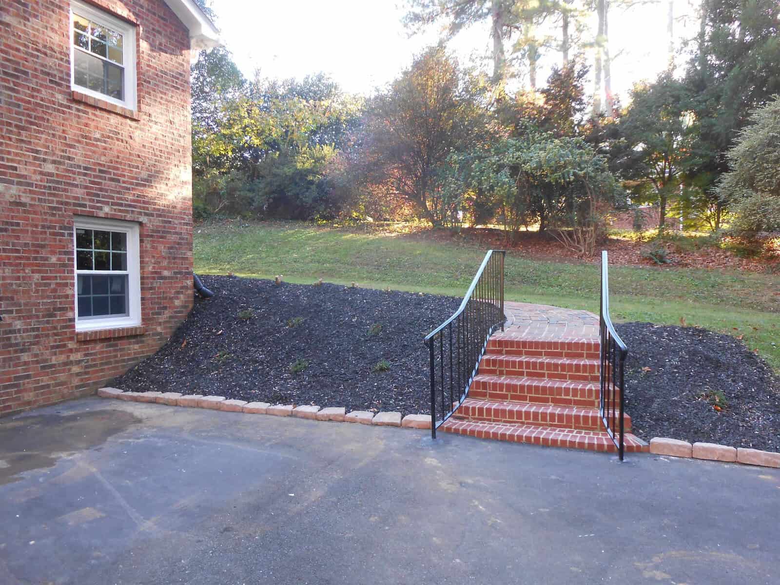 A brick house with a small window, adjacent to a newly mulched area leading to red brick steps with a black metal railing, surrounded by trees in a suburban setting.