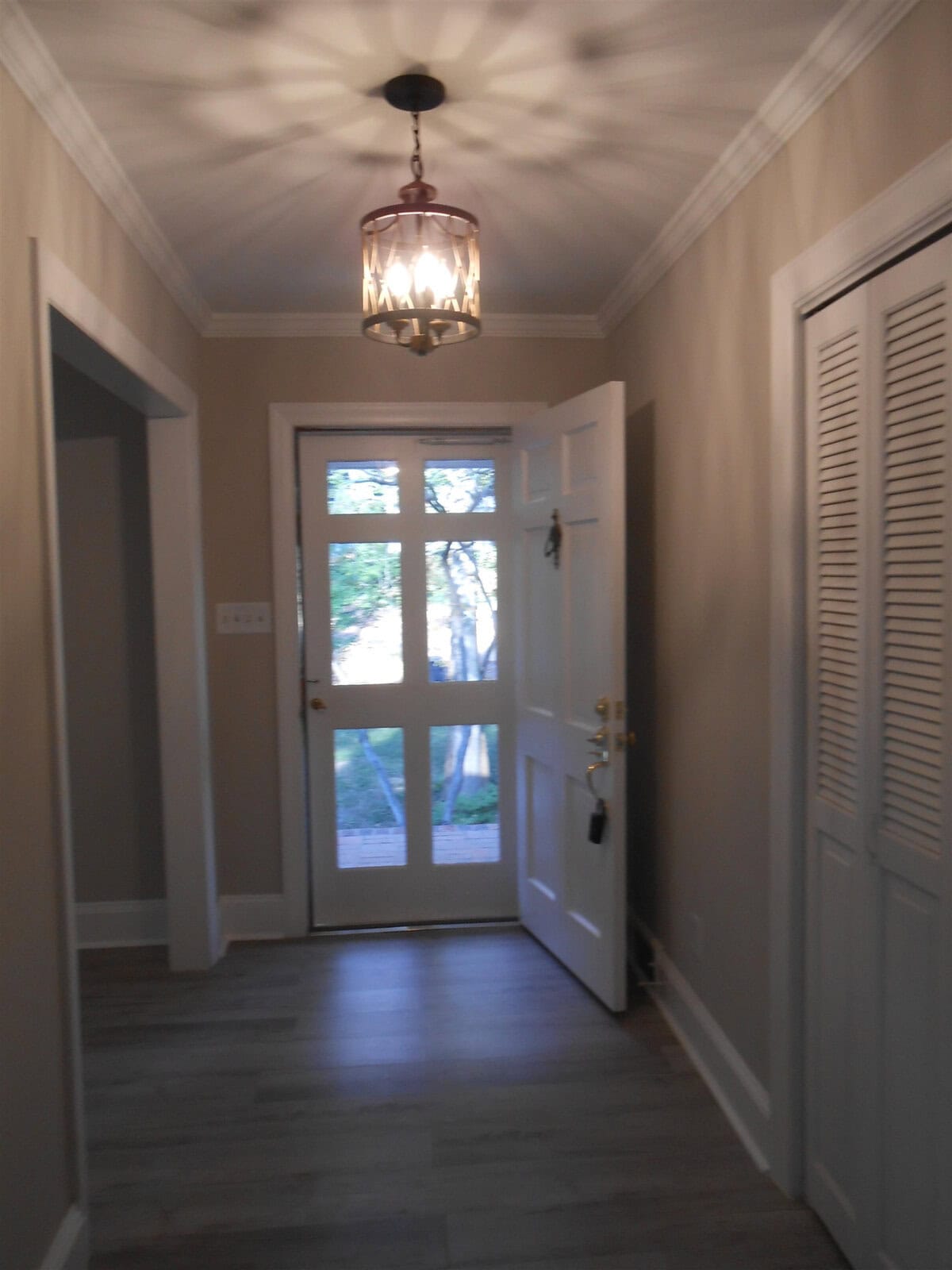 An interior view of a hallway with beige walls, a wooden floor, and a hanging chandelier. the hallway leads to a glass door at the end, showing a glimpse of the outdoors.