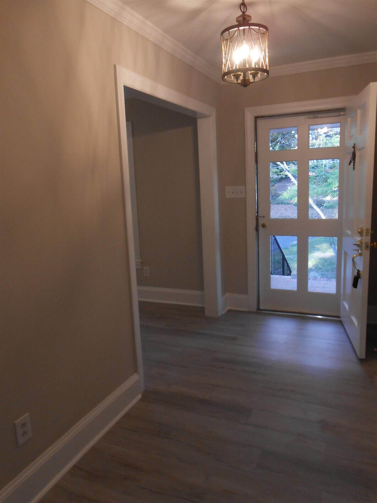 A brightly lit foyer with beige walls and gray wooden flooring, featuring a black front door with glass panels and a stylish hanging light fixture.