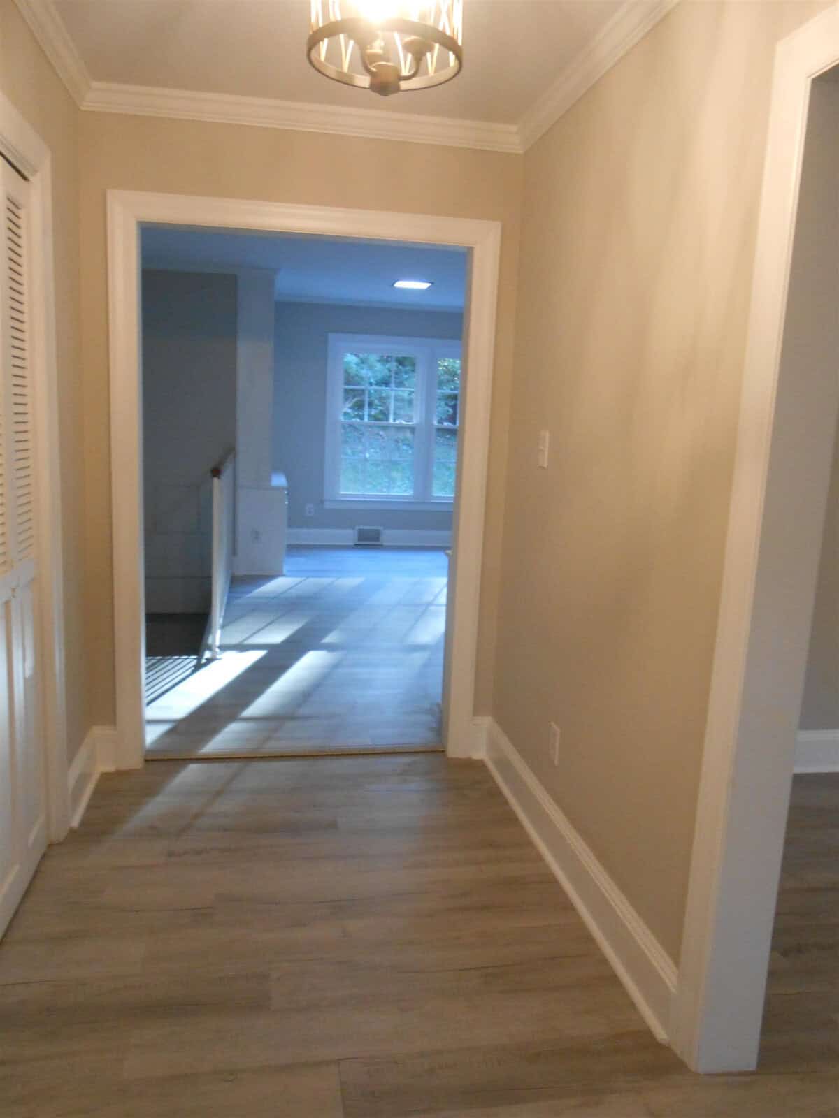 A hallway in a home with light wood floors and beige walls, leading towards a room with a blue wall visible through a doorway. a light fixture hangs from the ceiling.