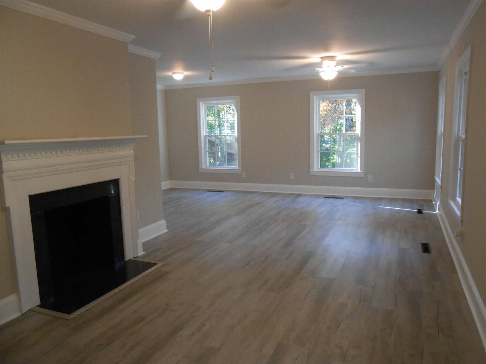 Spacious empty room with light gray walls, hardwood floors, a white fireplace, and two windows letting in natural light. the room has ceiling lights and is unfurnished.
