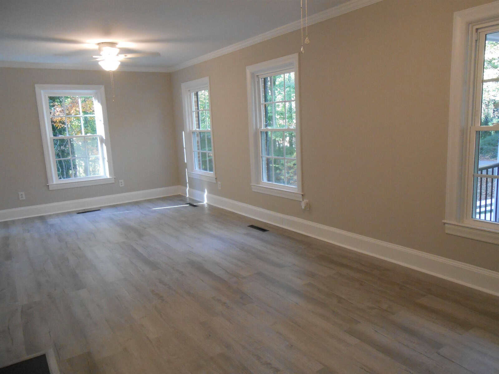 Empty living room with gray laminate flooring, off-white walls with white trim, and multiple windows letting in natural light, leading to a deck area outside.