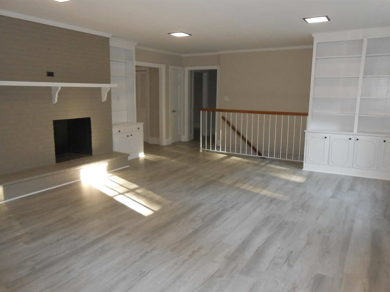 A spacious living room featuring a hardwood floor, a brick fireplace on the left, white built-in shelves, and an open doorway leading to another room. sunlight streams in creating patterns on the floor.