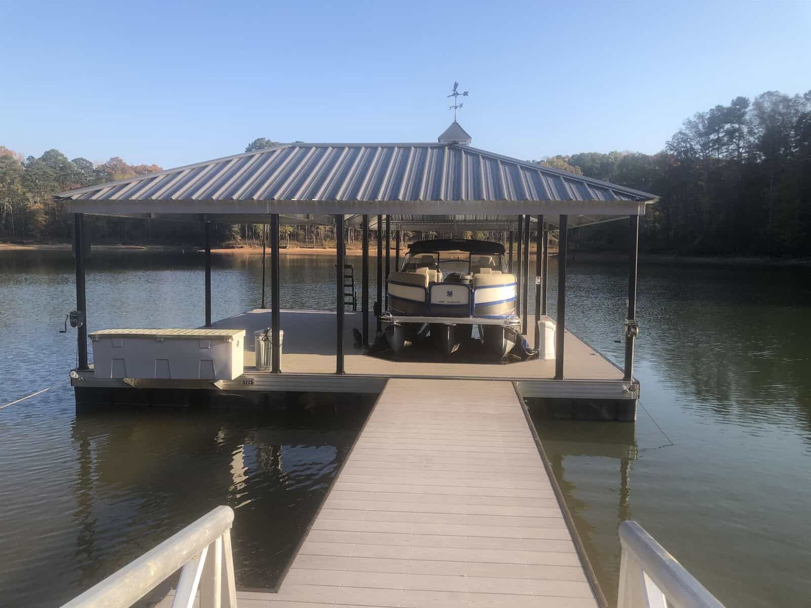A pontoon boat is docked under a metal roofed boat shelter at the end of a wooden walkway extending over a calm body of water in Henderson, NC.