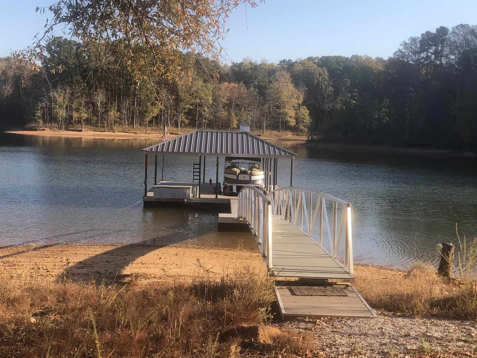 A boat docked under a covered slip on a tranquil lake with trees displaying autumn colors in the background.