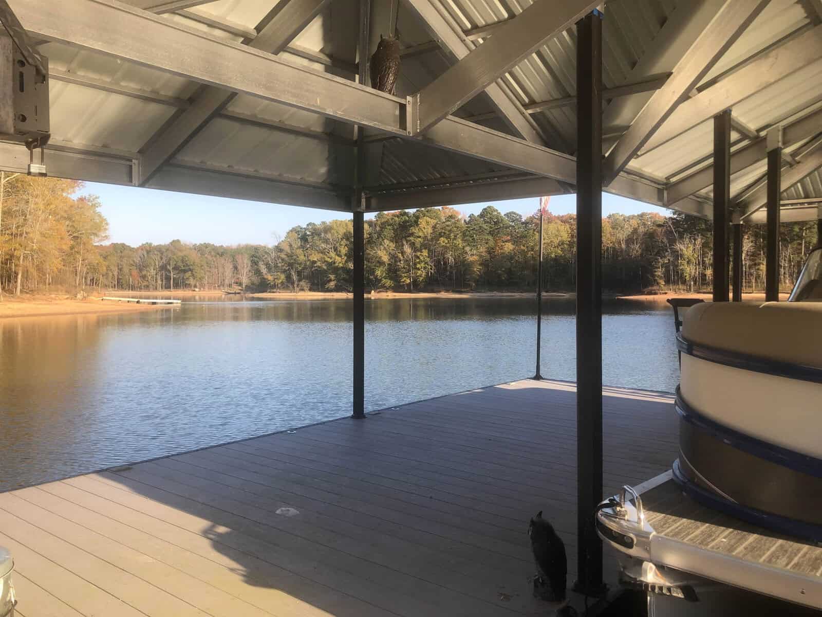 A covered wooden dock extends over a calm lake, with a pontoon boat to the right and a cat sitting on the edge, gazing out over the water. trees with autumn foliage are visible on the opposite shore under a clear sky.