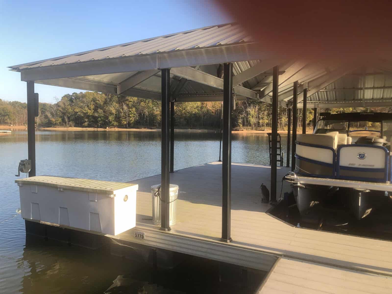 A covered boat dock on a calm lake with a pontoon boat parked on the right side and a small floating dock attached to it.