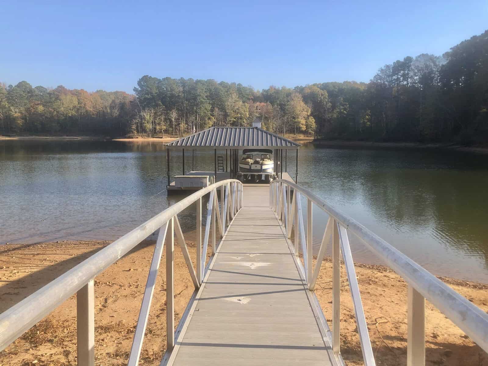 A metal walkway leads to a covered boat dock on a tranquil lake surrounded by autumnal trees.