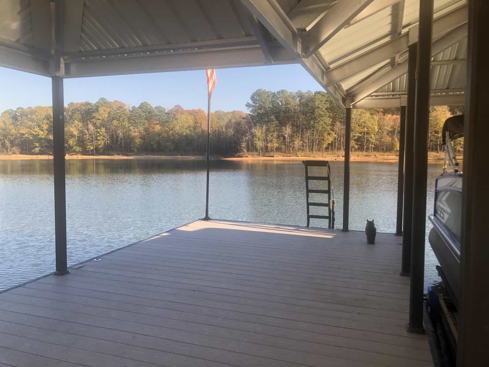A covered wooden dock overlooking a tranquil lake with a flagpole and an american flag to the left, a ladder leading into the water, and a small boat partially visible on the right.
