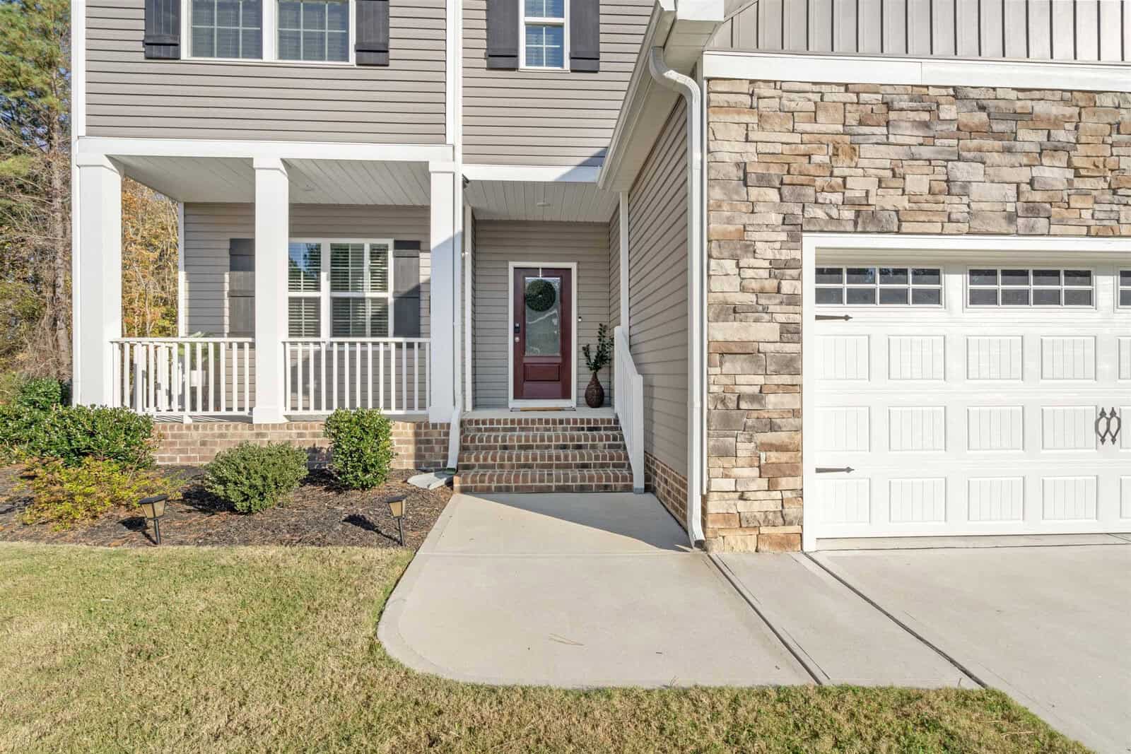 Front view of a house with a stone facade and white garage door. A covered porch with brick steps leads to a red front door. Manicured lawn and shrubs enhance the exterior.