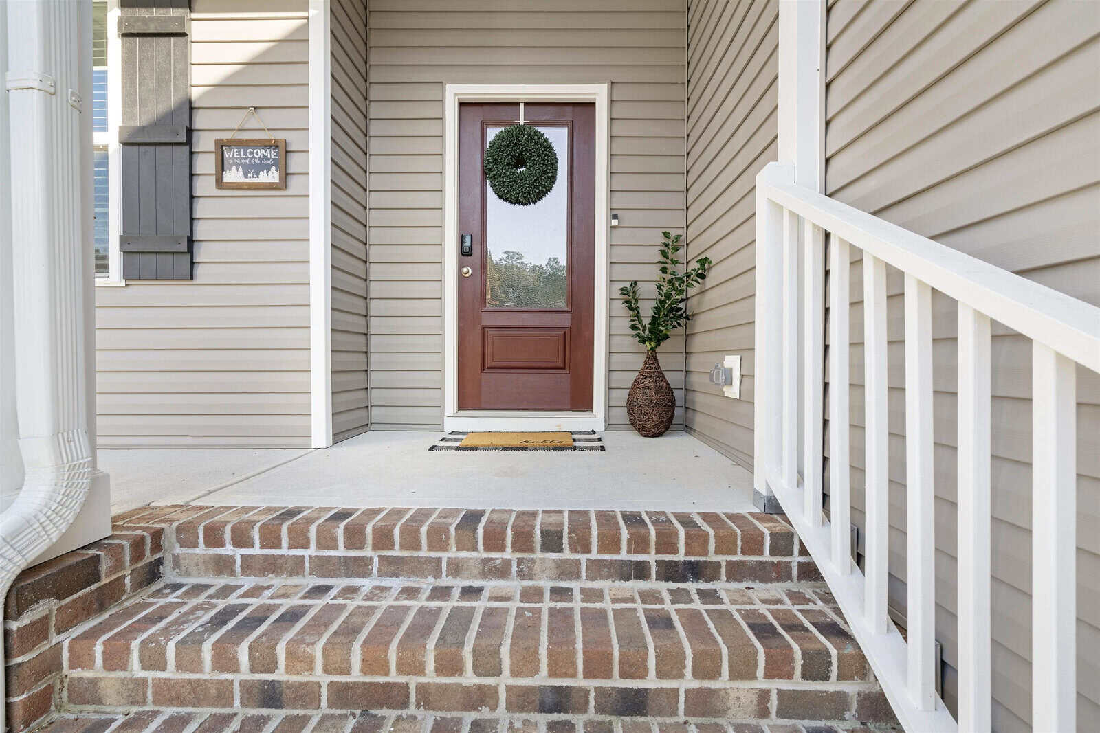 A front porch with brick steps leads to a brown door adorned with a green wreath. A welcome sign is on the siding, and a woven vase with greenery is beside the door. The area is framed by white railings and beige siding.