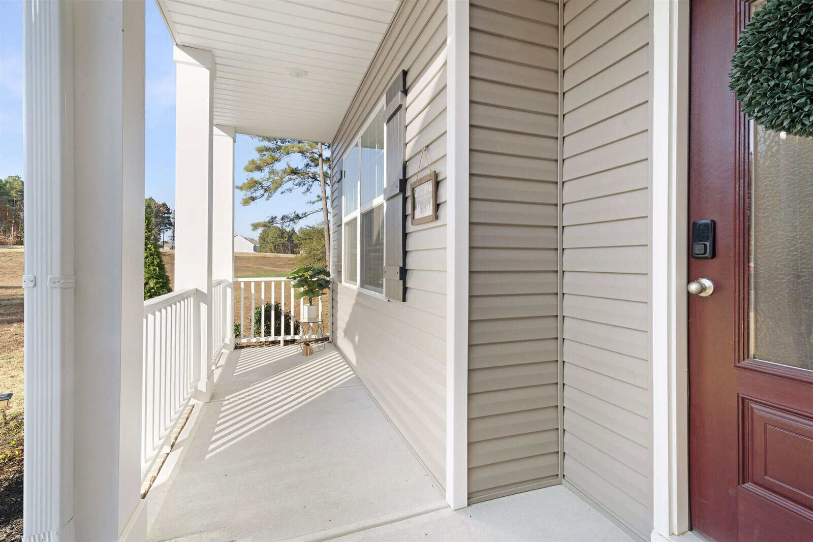 Front porch of a house with beige siding, a white railing, and a red door with a wreath. A small potted plant sits on the porch floor. The porch overlooks a grassy area with trees in the background.