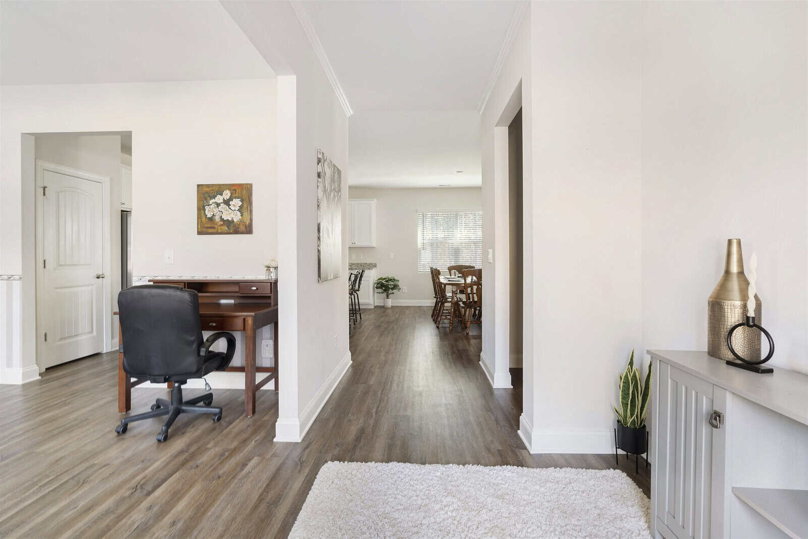A modern, open-concept interior features a home office with a black swivel chair and wooden desk to the left, leading to a dining area with a table and chairs in the background. Light wood flooring and white walls create a clean, airy atmosphere.