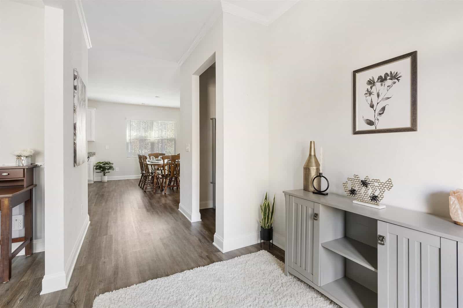 A bright hallway leads to a dining room with wooden chairs. The hallway features a cabinet with decorative items and a plant, a light-colored rug, and a framed floral picture on the wall. Hardwood floors run throughout the space.