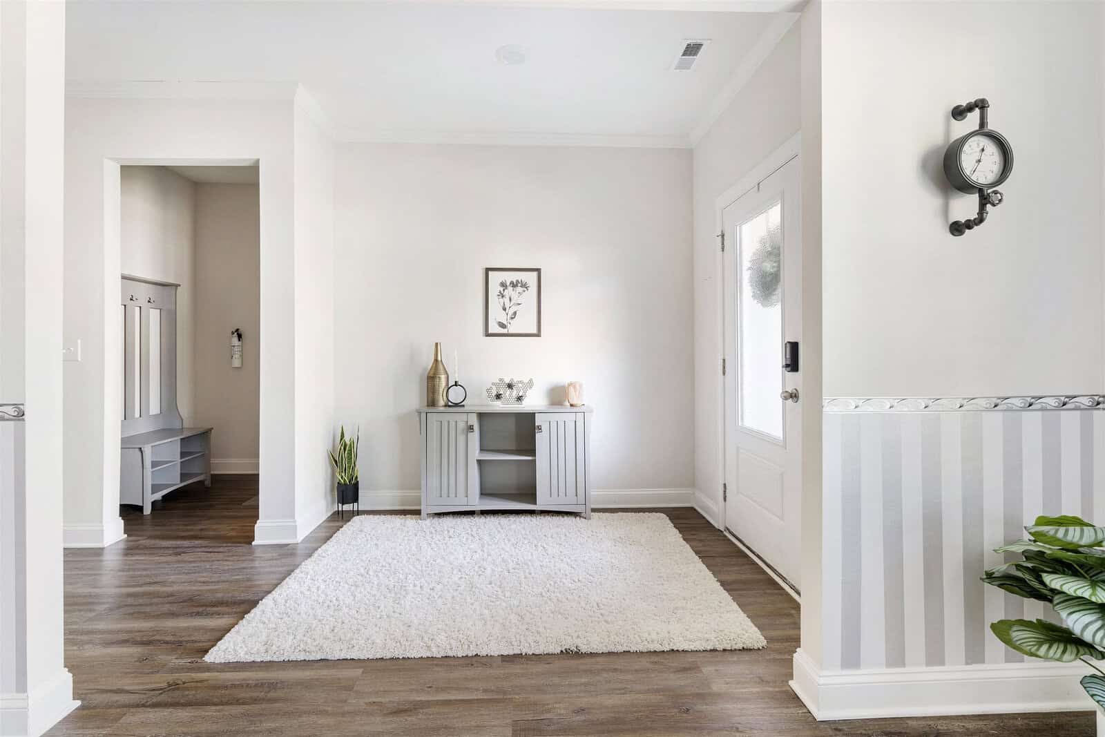 A bright, modern entryway with a light-colored console table on a white rug. The walls are white with striped wainscoting, and a small decorative clock hangs above. The room features light wood flooring and a plant in the corner.