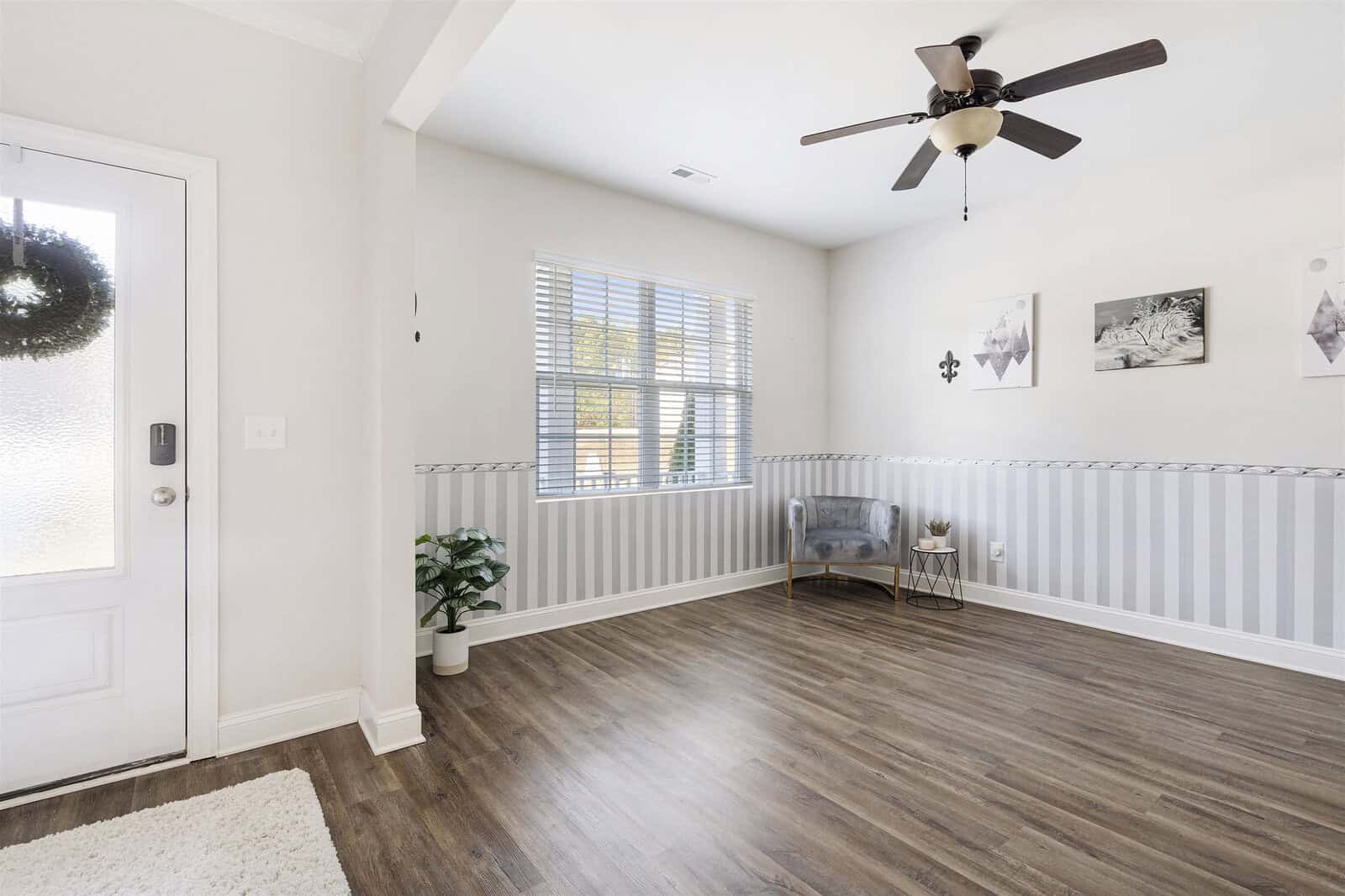 A bright, empty room with a wooden floor and white walls features a large window, ceiling fan, and wall art. A gray chair and small table with a plant decorate the corner, and a potted plant is near the entrance door with a wreath.