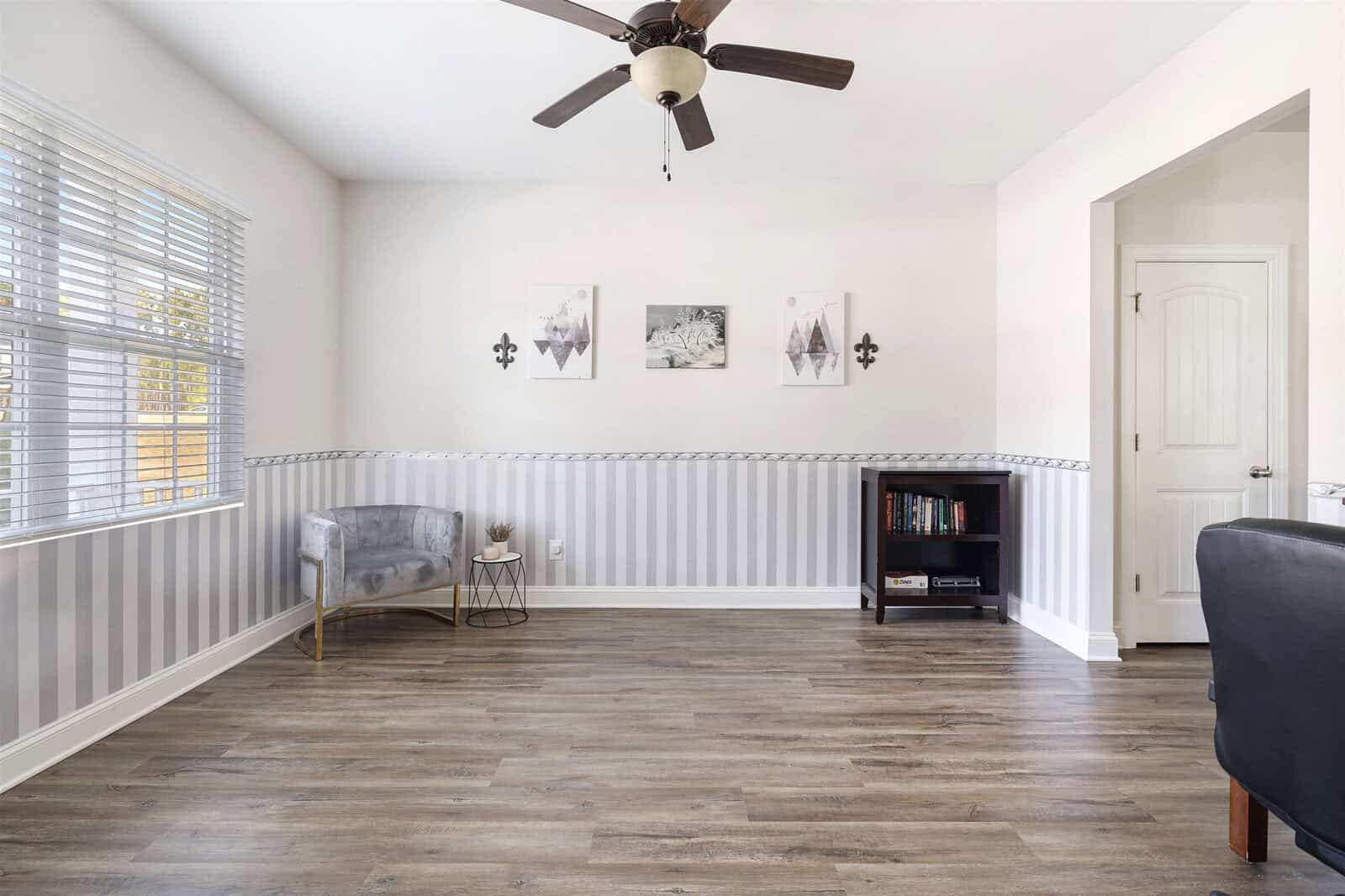 A bright, minimalist room with wooden floors, a ceiling fan, gray striped walls, two small framed artworks, a gray armchair with a side table, and a small bookshelf. A window lets in natural light, illuminating the space.