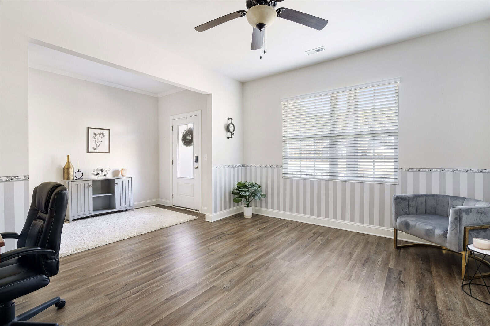 A bright, minimalist living room with wooden flooring features a ceiling fan, an office chair, and a gray armchair. Natural light streams in through a large window with blinds. A small entryway with a cabinet and decor is visible in the background.