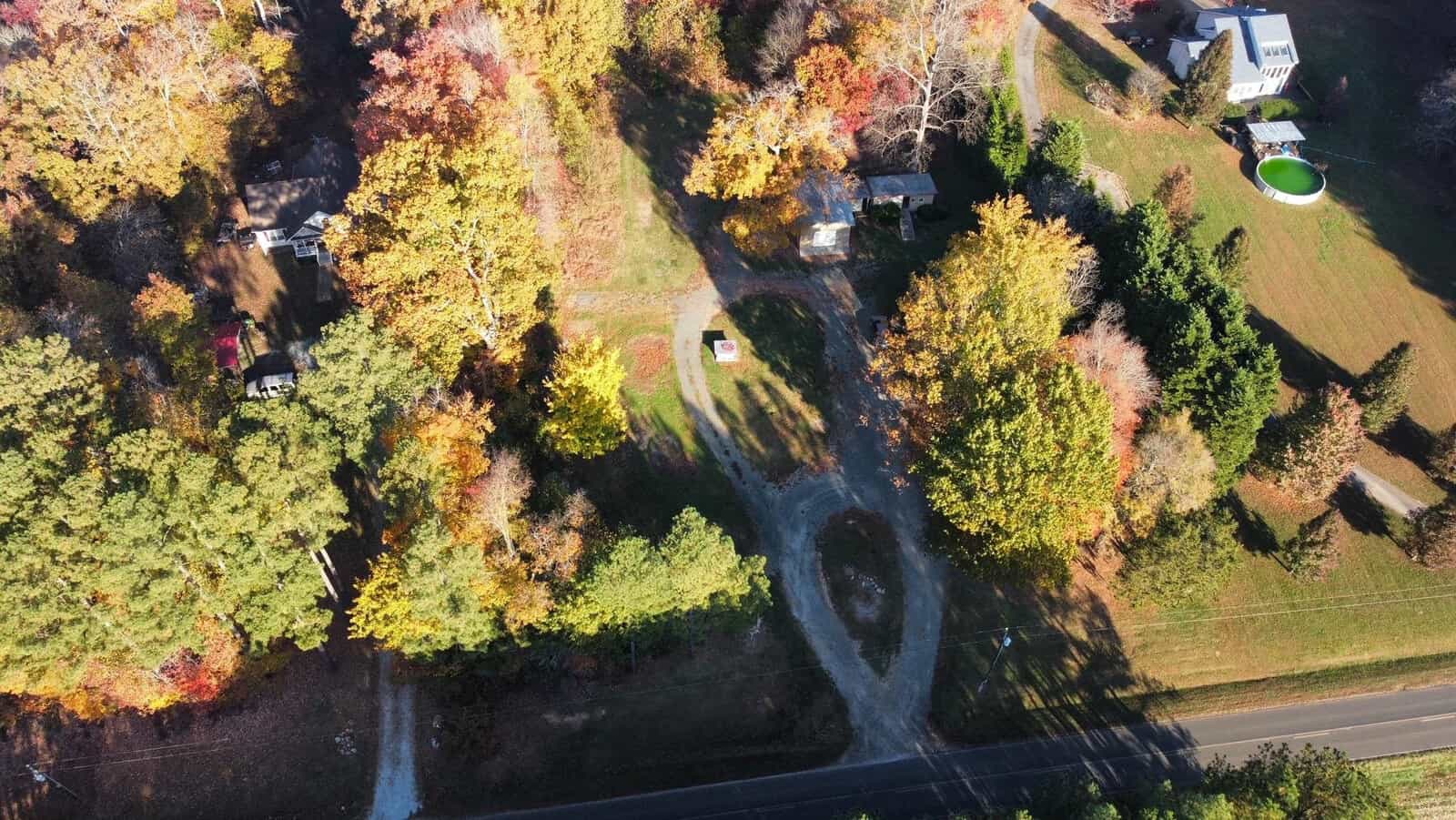 Aerial view of the rural landscape near 2037 Mountain Creek Road in Oxford, capturing autumn's vibrant palette. A tree-lined road meanders past scattered houses and a green pool, while the foliage in hues of orange, yellow, and red casts long shadows over the grassy terrain.