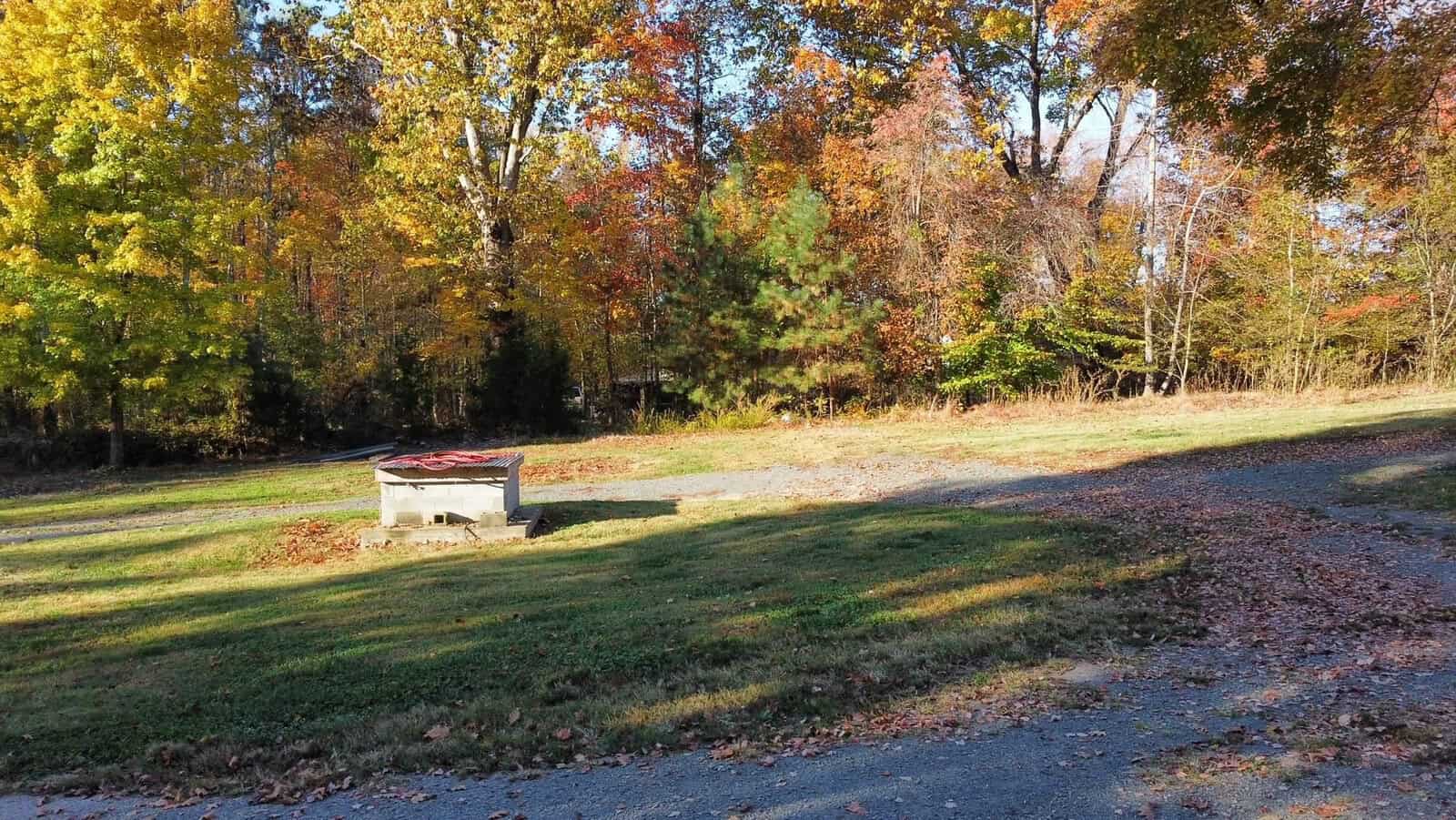 A small well cover stands in a grassy area surrounded by autumn foliage. Trees with colorful leaves in shades of yellow, orange, and red create a picturesque fall scene under a clear blue sky. A dirt path curves through the landscape.