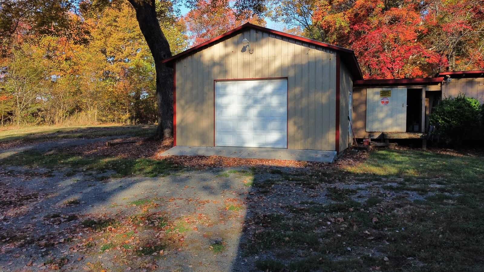 A small beige garage with a white door stands amidst autumn trees in vibrant shades of red, orange, and yellow. A driveway leads up to it, with fallen leaves scattered on the ground, casting shadows in the afternoon light.