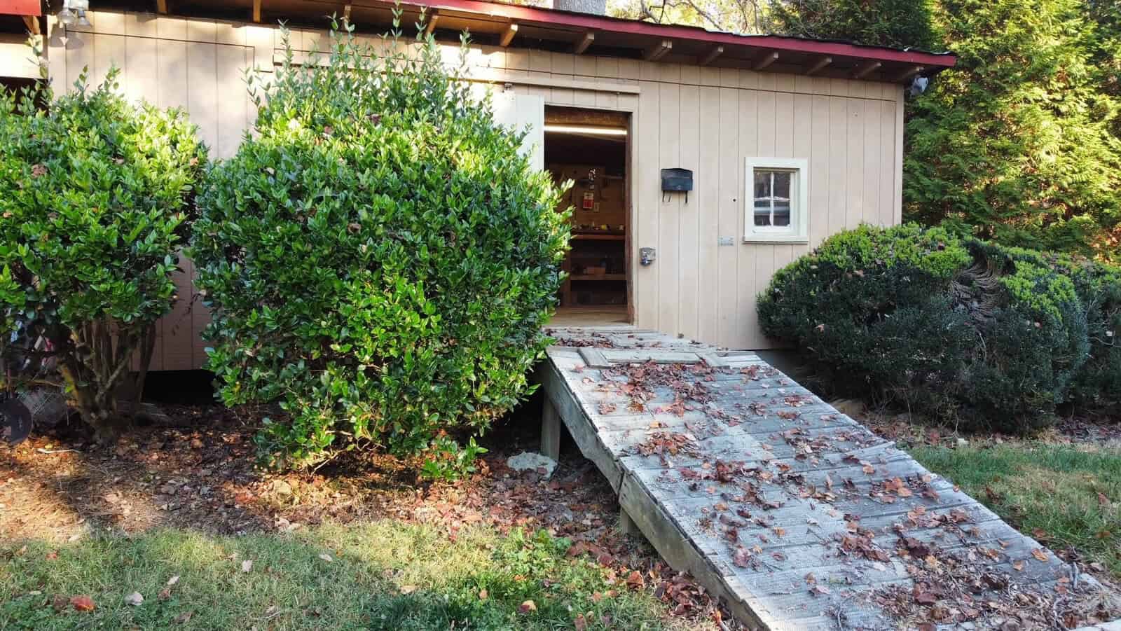 A small beige building with an open door, surrounded by bushes. A wooden ramp with scattered leaves leads to the entrance. The area is shaded by trees, and sunlight highlights the greenery.