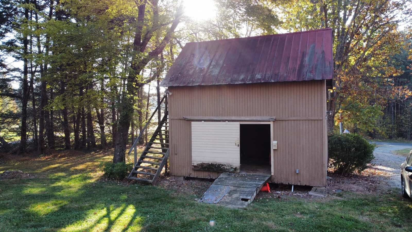 A rustic, two-story wooden shed with a weathered red roof is surrounded by trees. The sun shines through the leaves. A wooden staircase leads to an upper door, while a ramp and bushes are by the lower entrance. A gravel path is visible nearby.