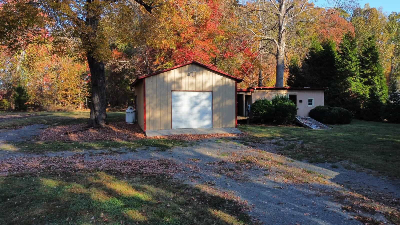 A small beige garage and a house are surrounded by vibrant autumn trees with red and orange leaves. The ground is partially covered with fallen leaves. A gravel driveway leads up to the garage.