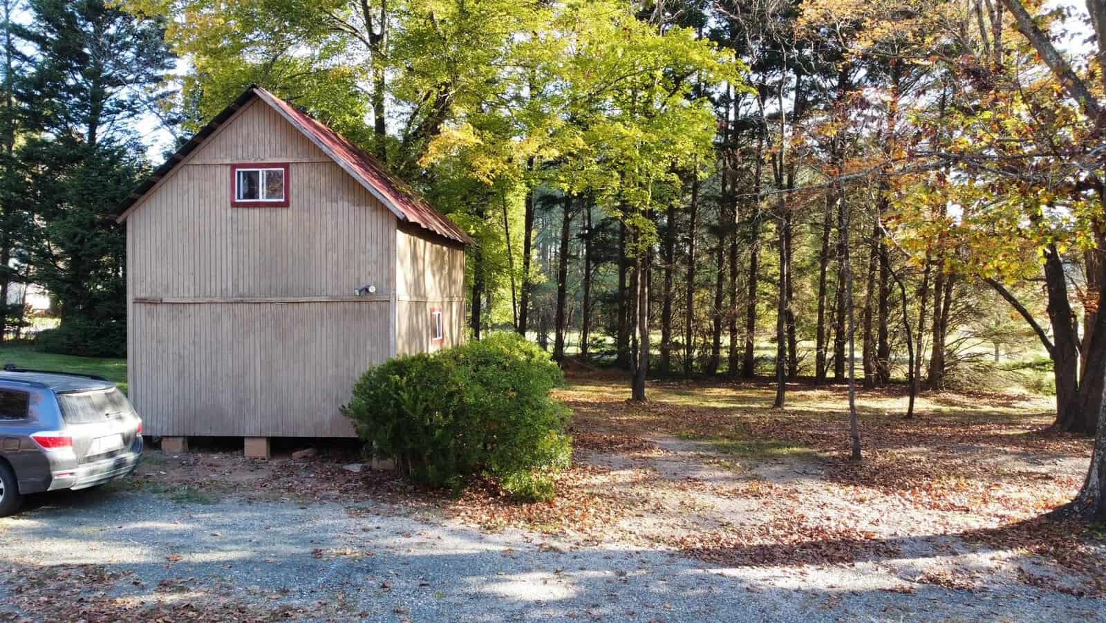 A small wooden cabin with a pitched roof sits next to a parked car amidst a forested area. The ground is covered with fallen autumn leaves, and tall trees with green and yellow foliage surround the cabin, creating a serene, natural setting.