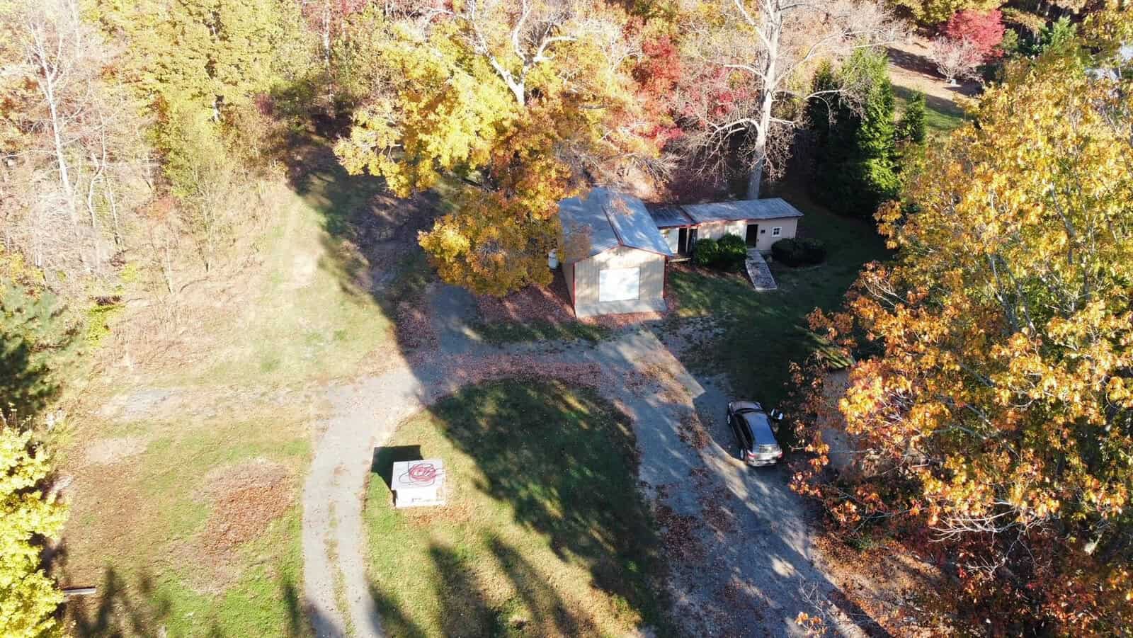 Aerial view of a rural area with a small white house surrounded by autumn trees with yellow and orange leaves. A car is parked on a dirt driveway nearby. A small sign is visible next to the driveway.