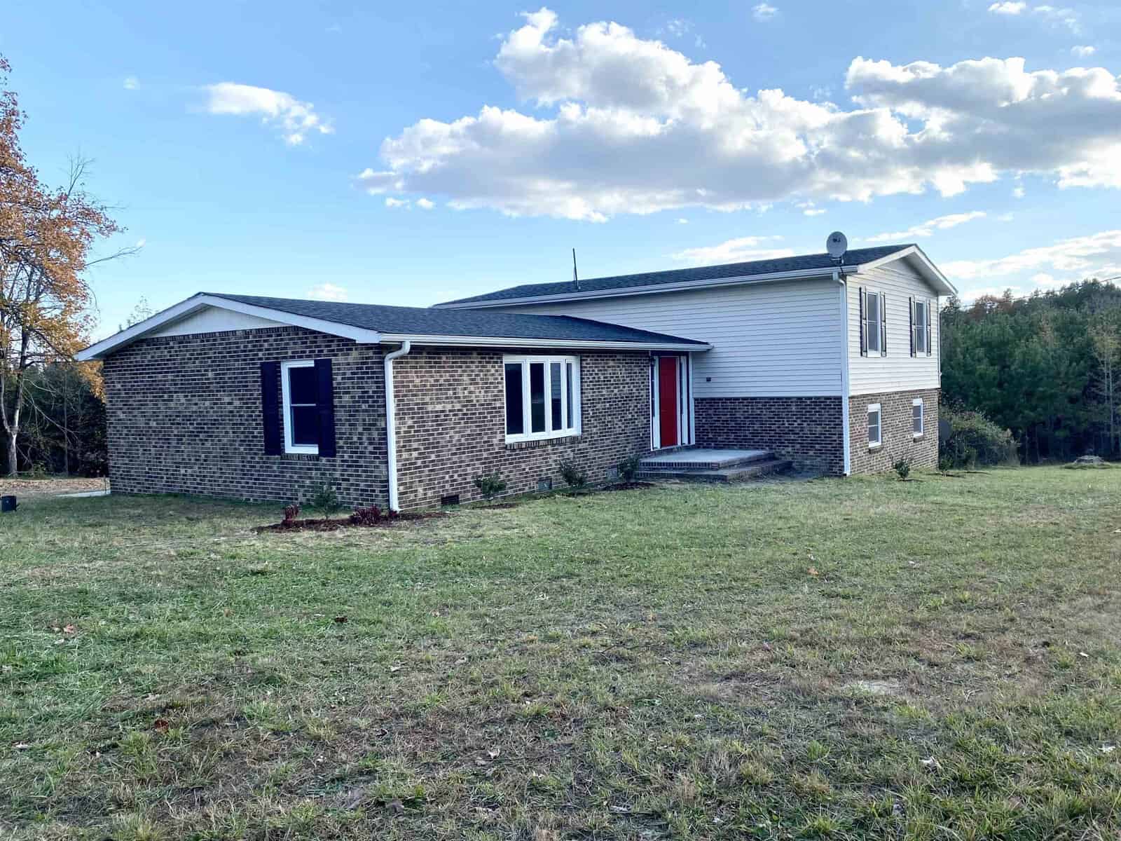 A split-level house with brick and siding exteriors, featuring a red front door, surrounded by a grassy yard and trees under a clear, partly cloudy sky.
