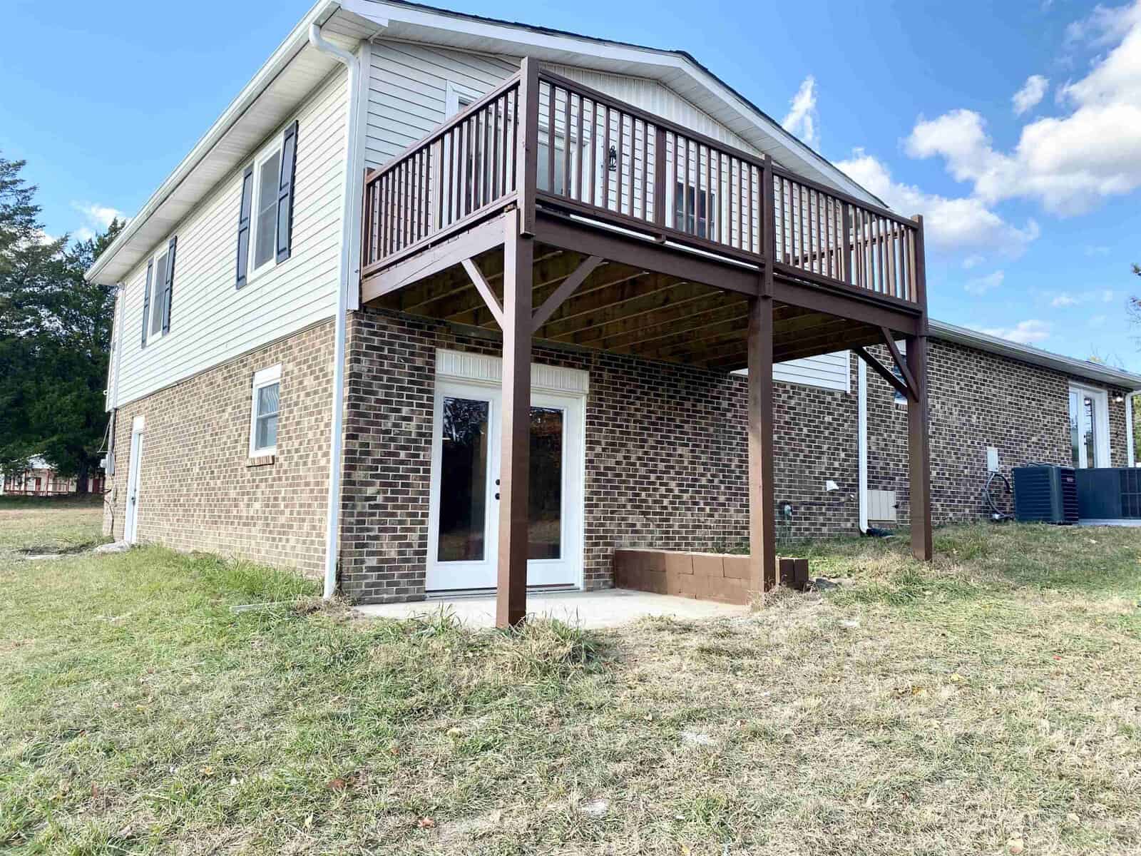 A two-story brick house featuring a raised wooden deck accessed by stairs, surrounded by a grassy yard under a clear blue sky.