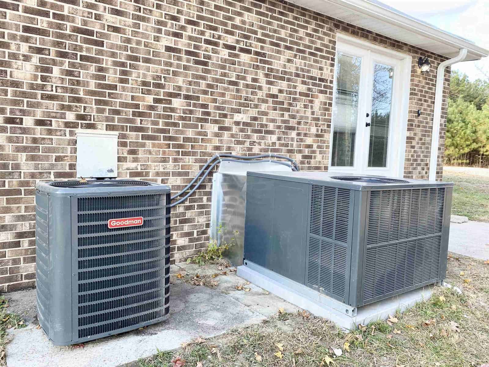 Two large air conditioning units are installed outside a brick house next to a bay window, with cables running from the units to the house.