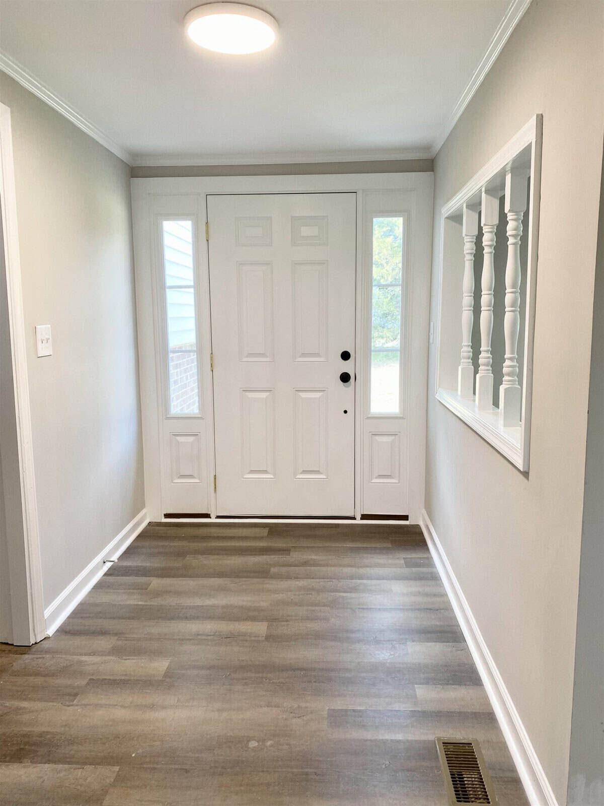 A clean, well-lit hallway with wooden flooring leading to a white door, flanked on the left by a row of decorative wall-mounted white columns and a rectangle mirror on the right.