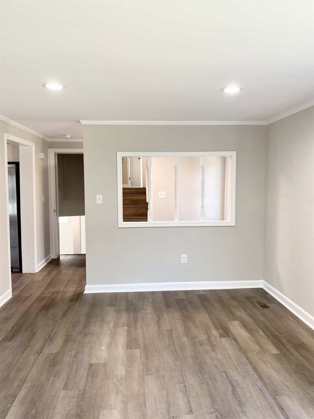 Interior view of an empty room with light gray walls, wooden flooring, and a square interior window opening towards another room. bright natural light fills the space.