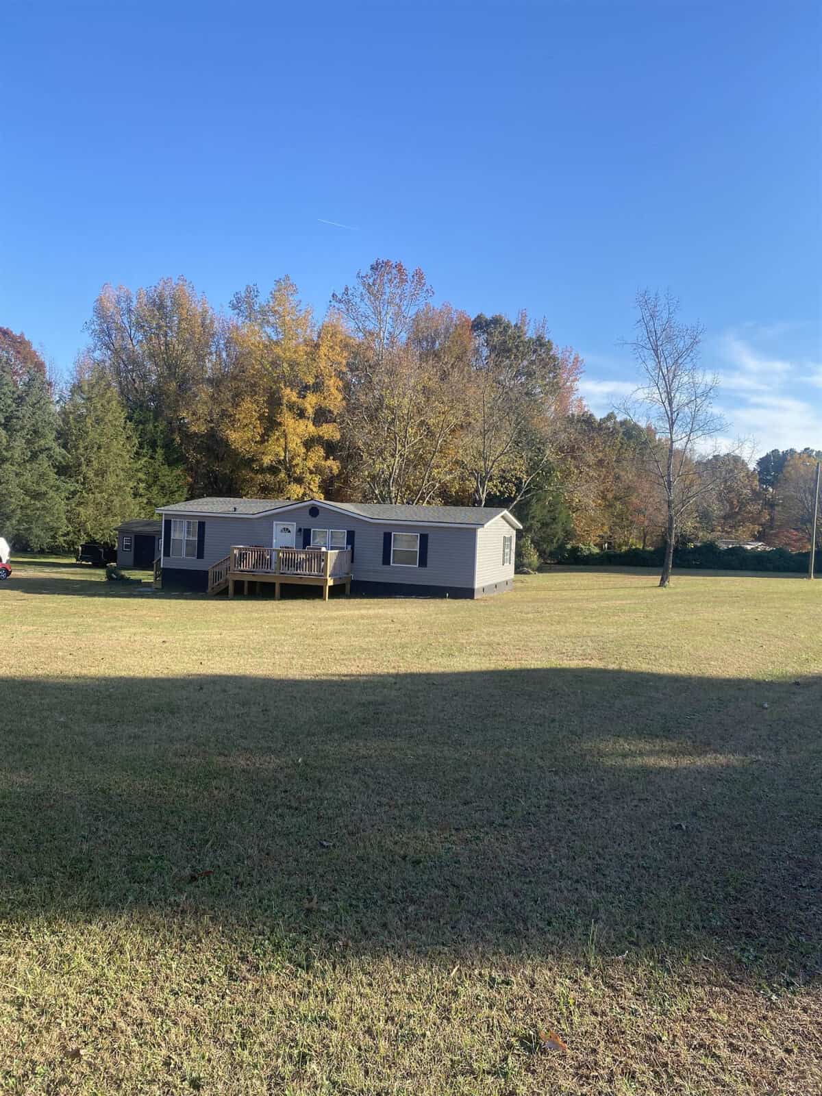 A gray mobile home with a small wooden porch sits on a large grassy yard. The background features trees with autumn foliage under a clear blue sky.