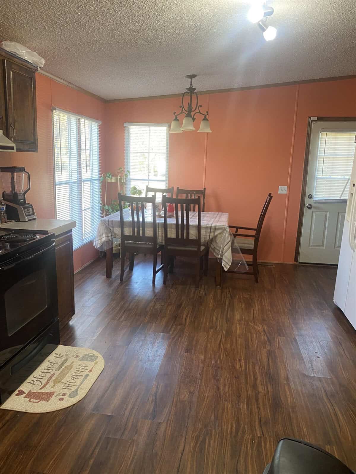 A cozy kitchen with orange walls and wooden flooring. A dining table with a checkered tablecloth and six chairs is situated by the windows. A light fixture hangs above the table, and a "Bless this Home" mat is near the stove.