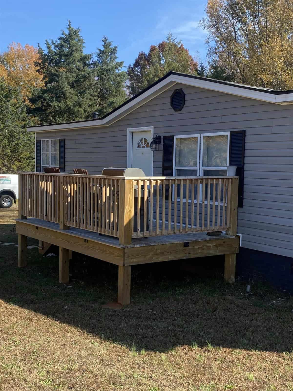 A small mobile home with beige siding and dark shutters, featuring a small wooden deck with a railing in front. It is set on a grassy lawn with trees and a vehicle visible in the background.