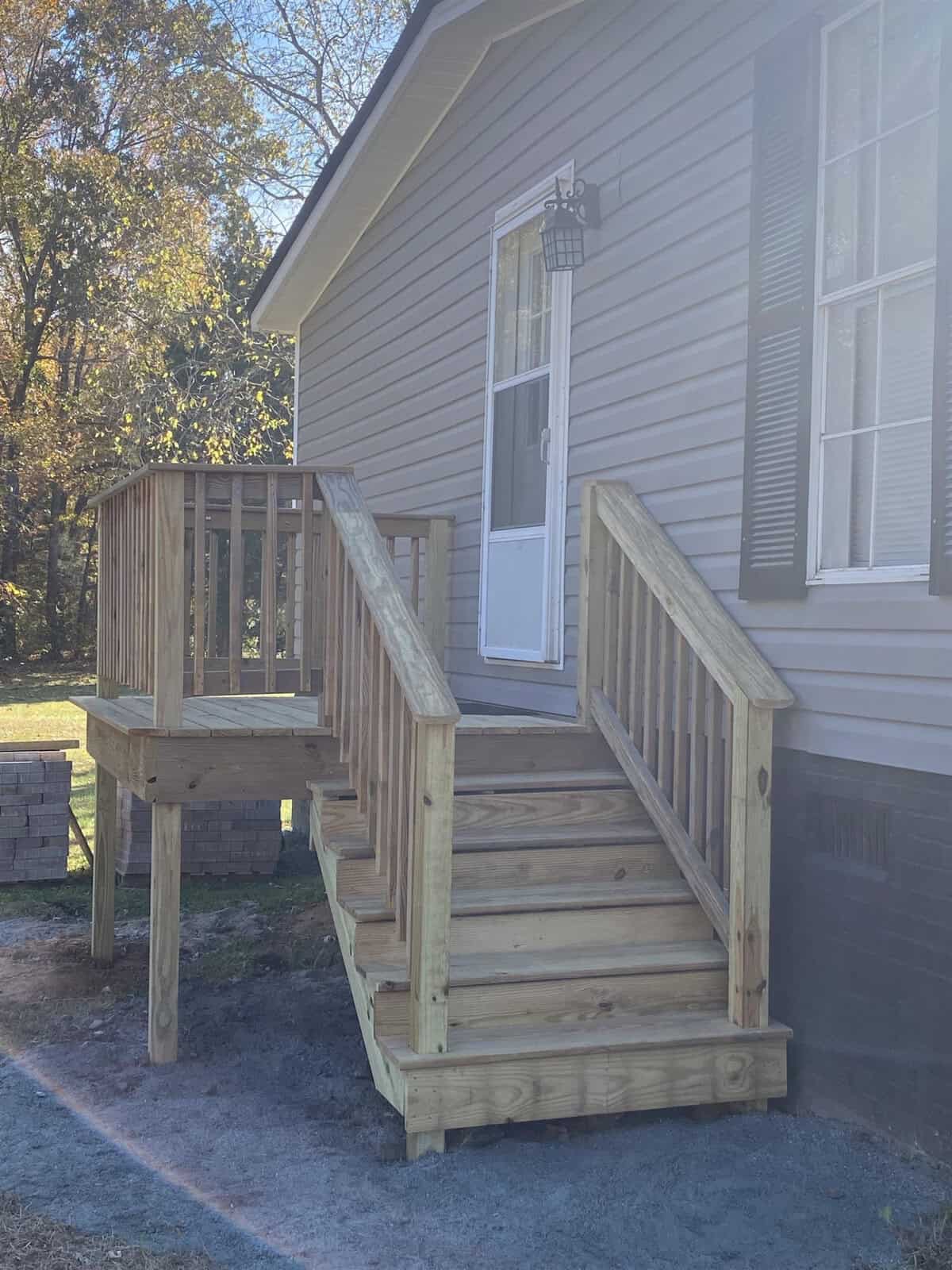 A small wooden porch with stairs leads to the side door of a house. The house has beige siding and a single window with dark shutters. There's a lantern style light fixture by the door. Trees and brickwork are visible in the background.