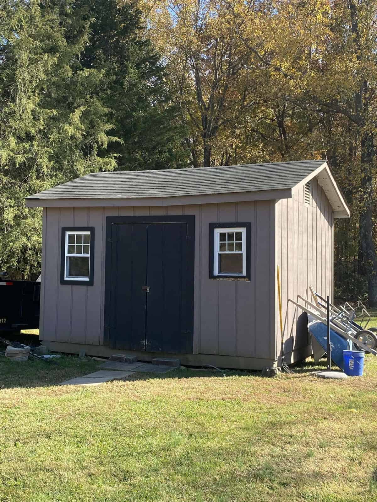 A small wooden shed with two windows and a dark double door, surrounded by grass. A wheelbarrow and a blue bucket sit to the right. Tall trees with autumn foliage are visible in the background.