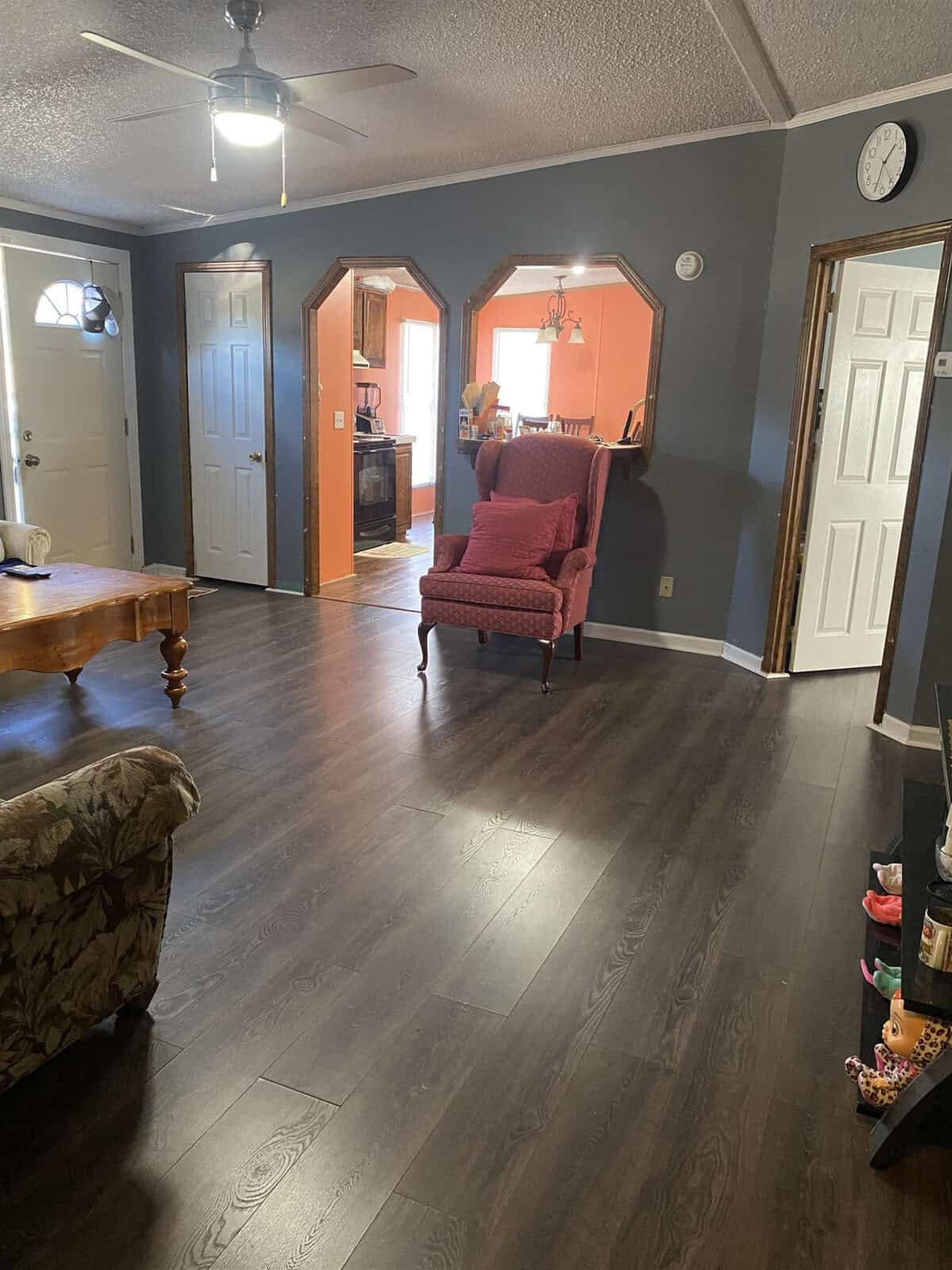 A cozy living room with dark wood flooring, featuring a pink armchair and a wooden coffee table. The walls are painted dark gray. Three doorways lead to other rooms, one glimpse showing a warm-toned dining area. A ceiling fan is on and a clock is mounted high.